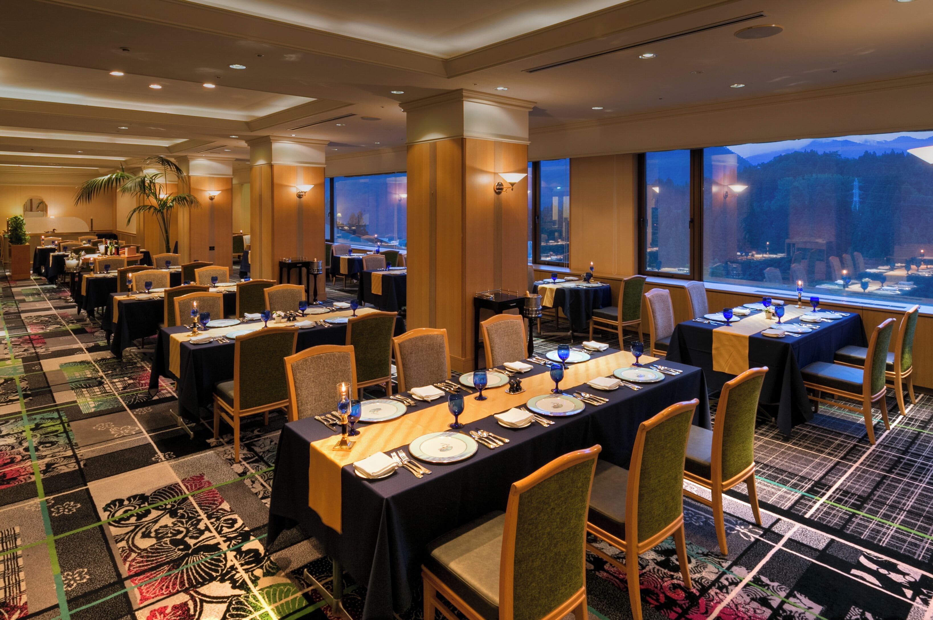 A banquet hall with tables set for dining, large windows with a view of a cityscape, and a patterned carpet.