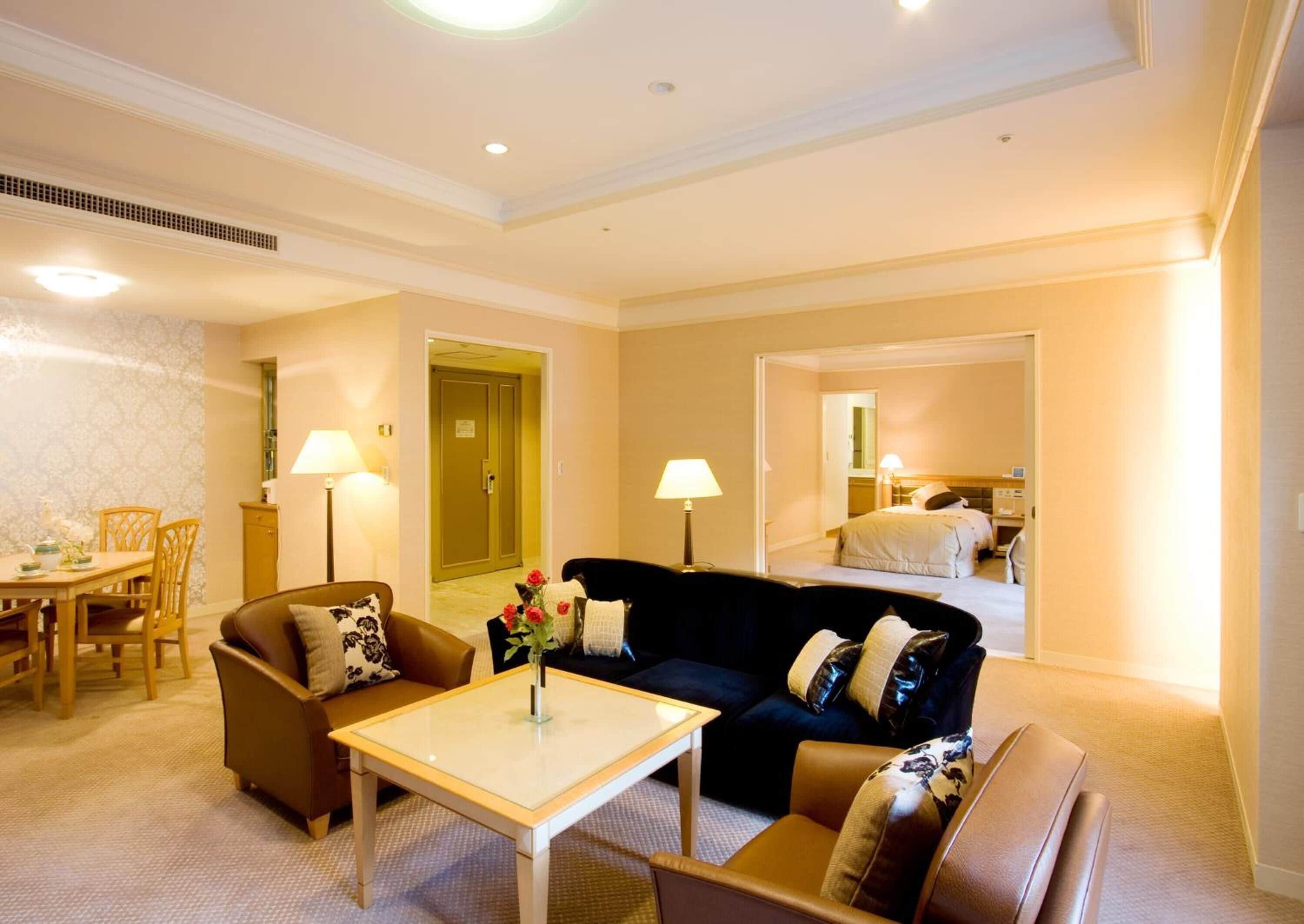 Habitación de hotel con sala de estar que incluye un sofá, una mesa de centro y una mesa de comedor con sillas. Se ve una puerta que da a un dormitorio con cama y un baño al fondo.