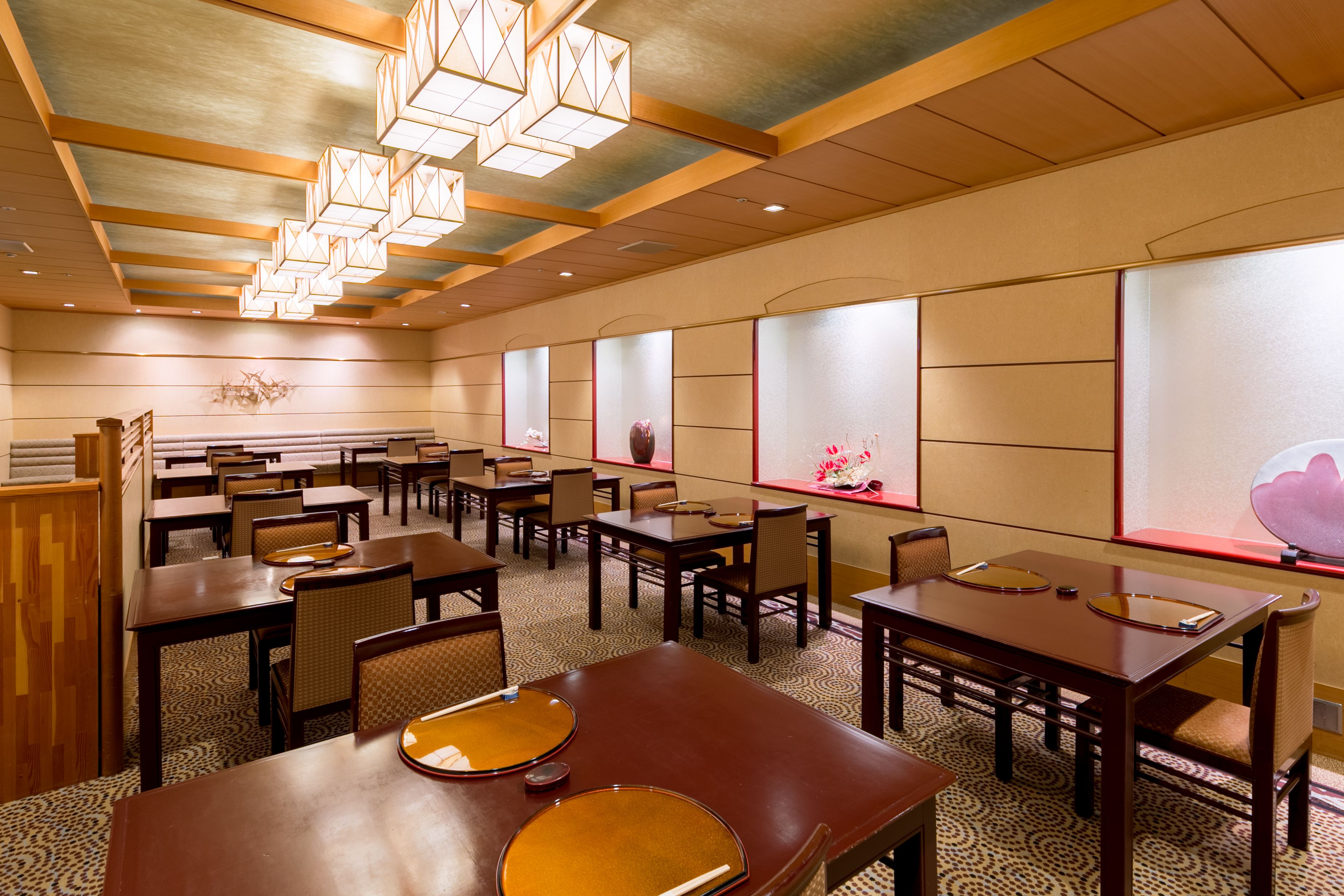 A hotel dining area with multiple tables and chairs, wooden ceiling with hanging lights, and large windows with decorative elements.