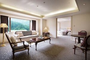 Amplia habitación de hotel con sofá, sillón y mesa de comedor. Un gran ventanal ofrece vistas a un área boscosa.