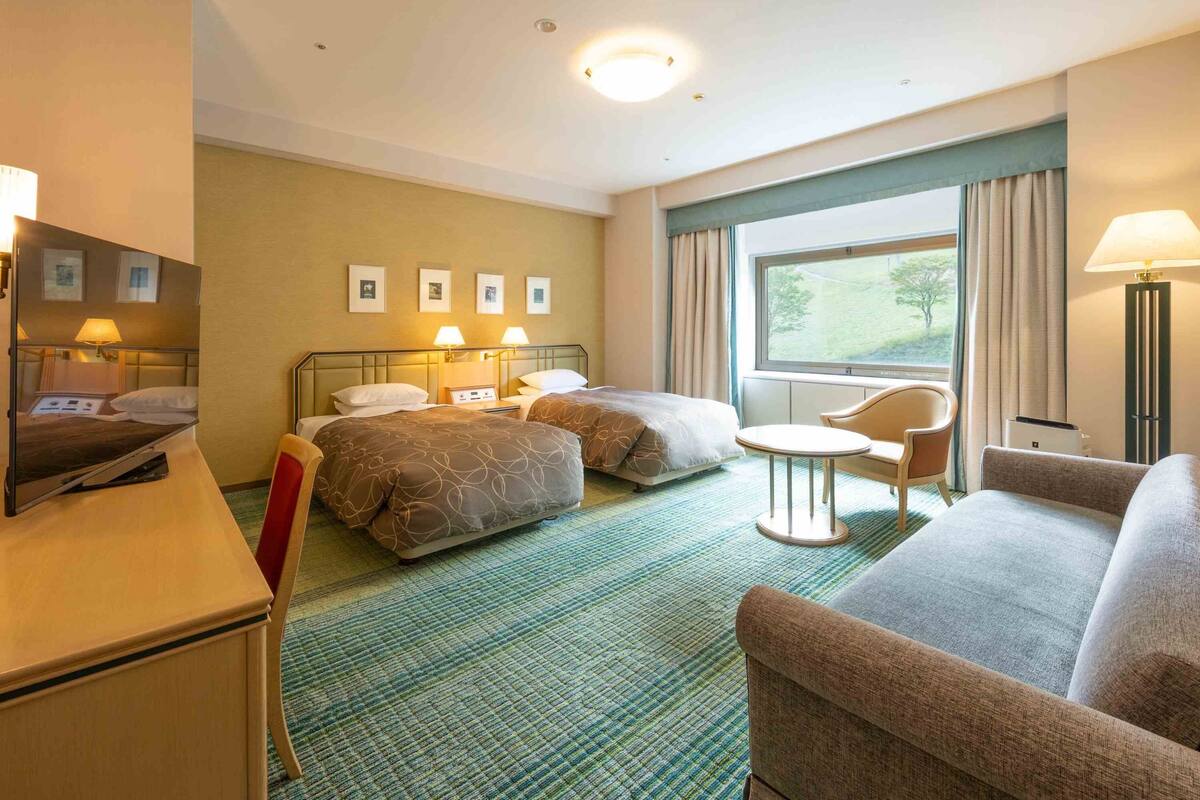 Une chambre d’hôtel avec deux lits, un bureau, une chaise, une petite table et une fenêtre donnant sur un paysage verdoyant.
