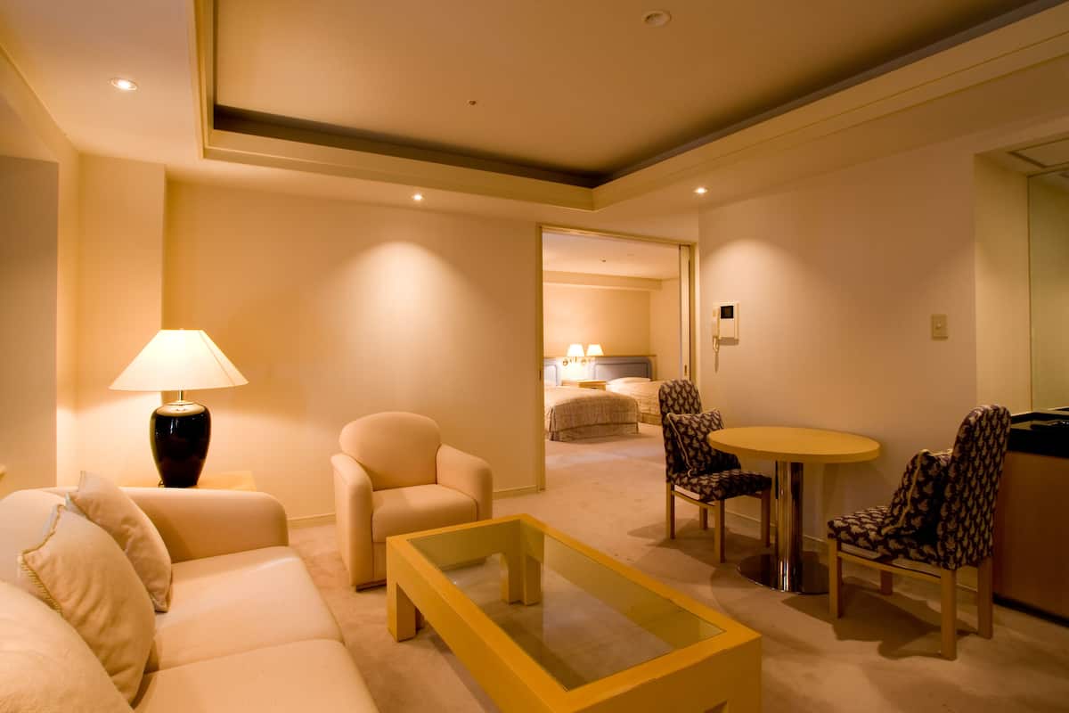 Une chambre d’hôtel avec un canapé, une table basse, une lampe, une table à manger avec des chaises, et un lit en arrière-plan.