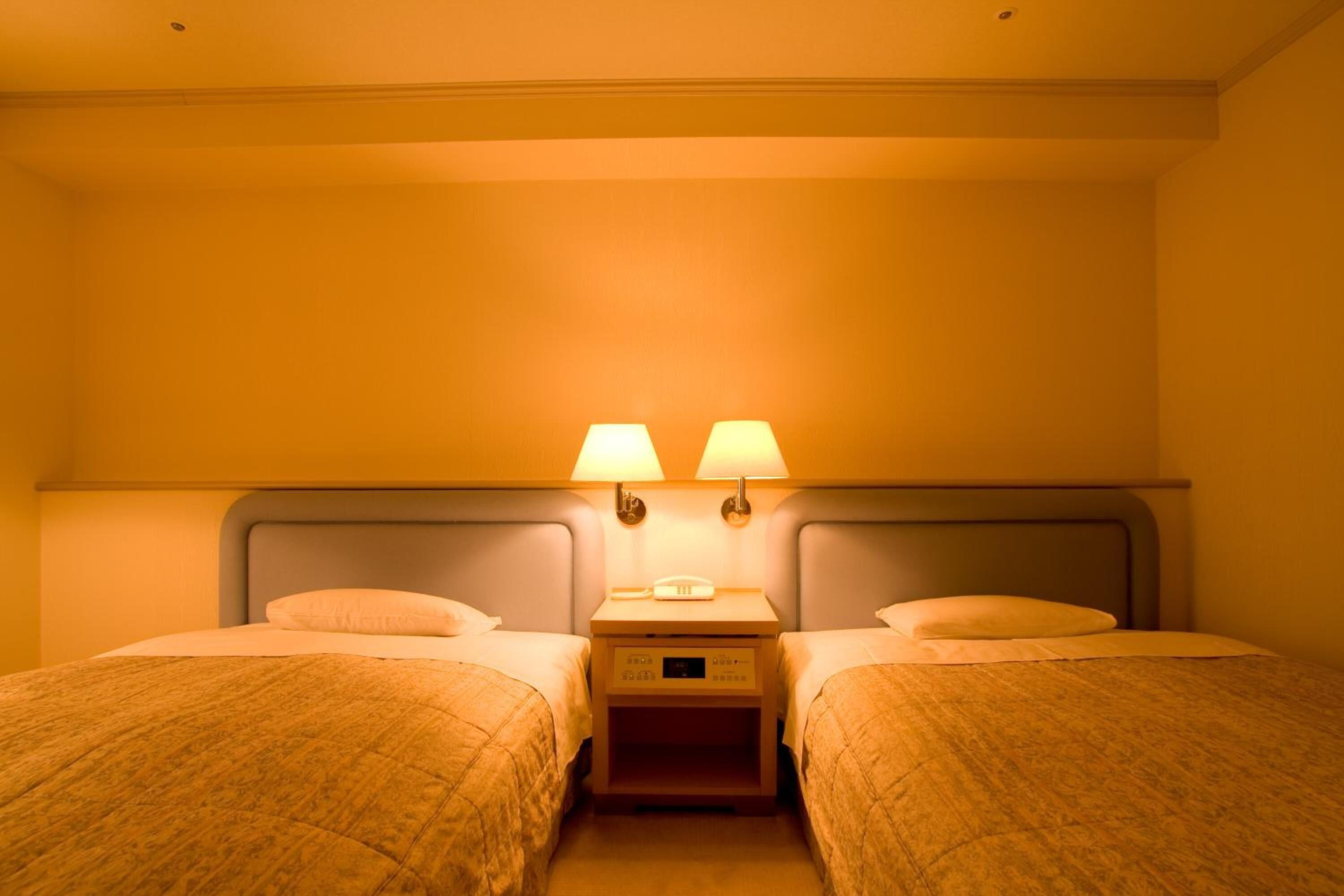A hotel room with two single beds, a bedside table with a lamp, and a wall-mounted light fixture.