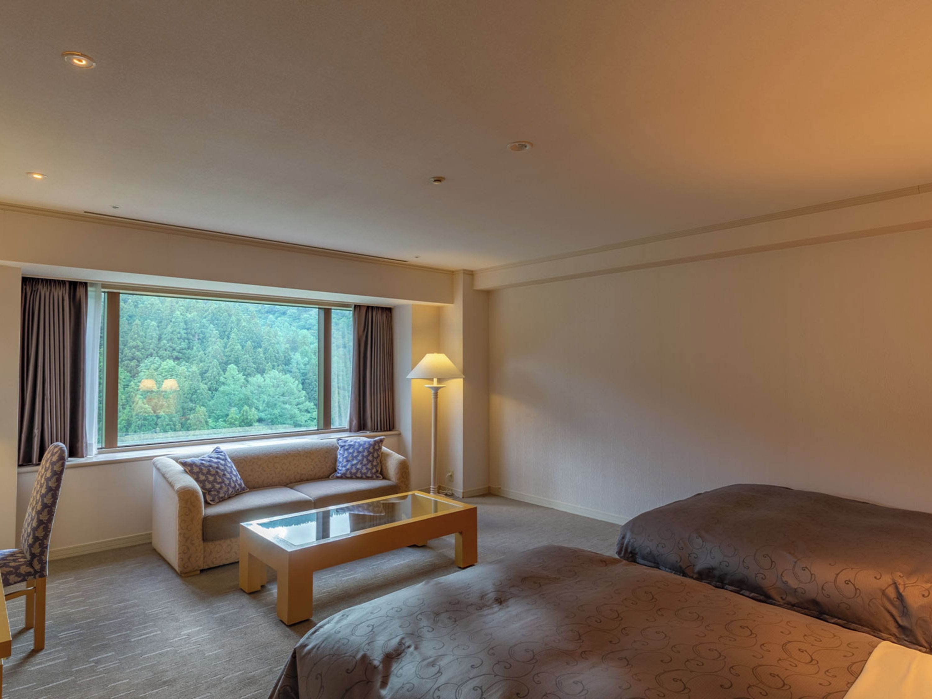 A hotel room with a sofa, a glass-top coffee table, a bed, and a window with curtains.