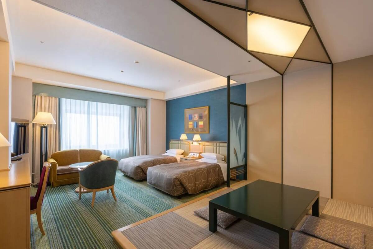 Une chambre d’hôtel avec deux lits, un bureau, une chaise, une table basse et une grande fenêtre avec des rideaux.