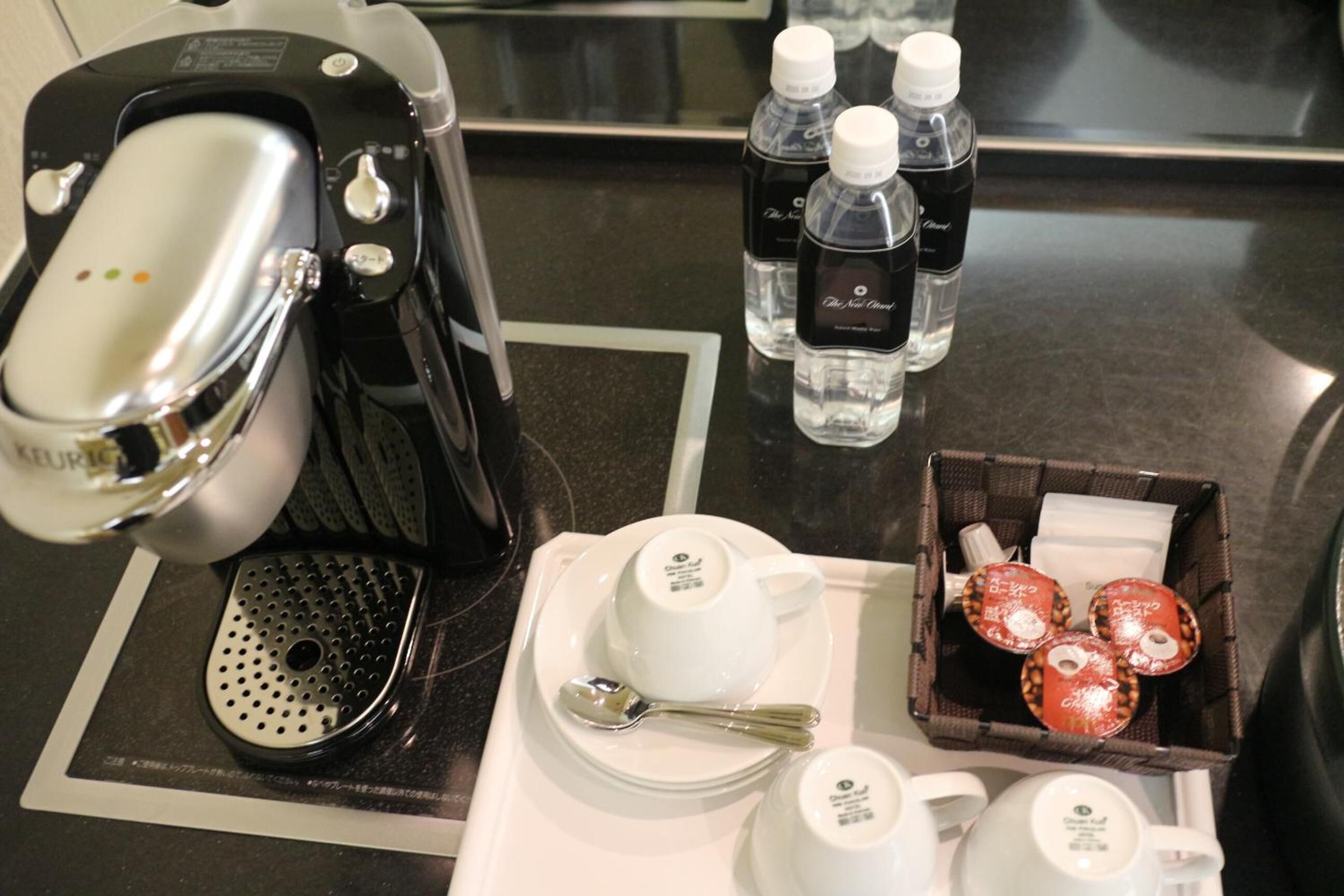 Preparación para el desayuno en una habitación de hotel, con cafetera, botellas de agua, tazas de té, una cuchara y una cesta de pasteles.