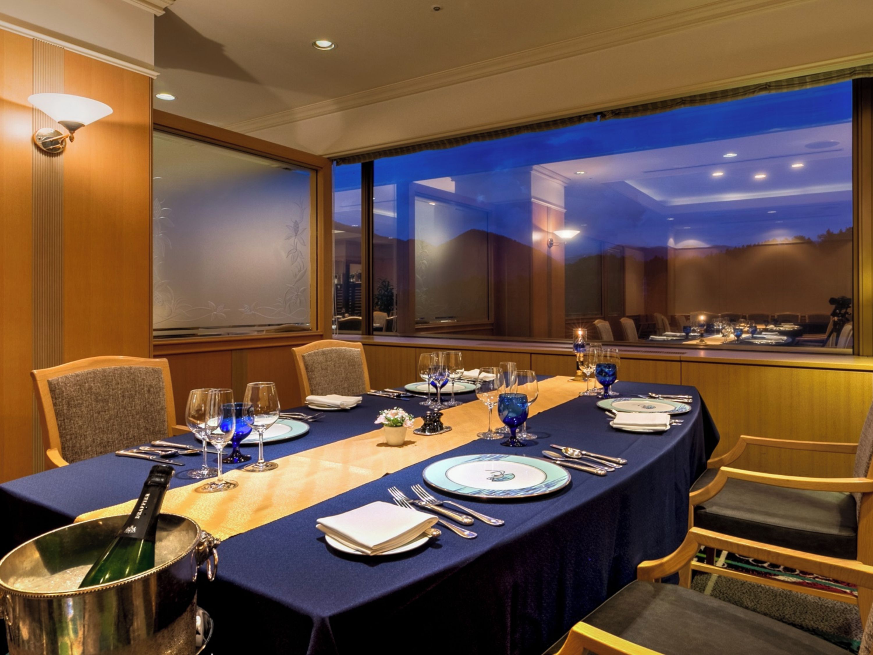A conference room with a long table set for a meeting, equipped with chairs, a projector, and a large window offering a view of the cityscape.