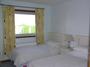 A bedroom with two single beds, a window with yellow curtains, a bedside lamp, and a view of a green field. - The Antlers (Lybster)