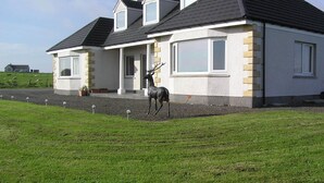 A two-story house with a stone facade, a black roof, and a deer statue in the front yard. - The Antlers (Lybster)