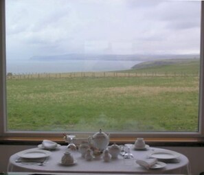 A table set with fine china and teapots, overlooking a grassy field and a distant view of a body of water. - The Antlers (Lybster)