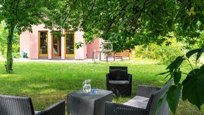 An outdoor seating area with wicker furniture and a table, a building with a glass door, and a garden in the background. - Roso 3 bedroom modern country house (gmina Chynów)