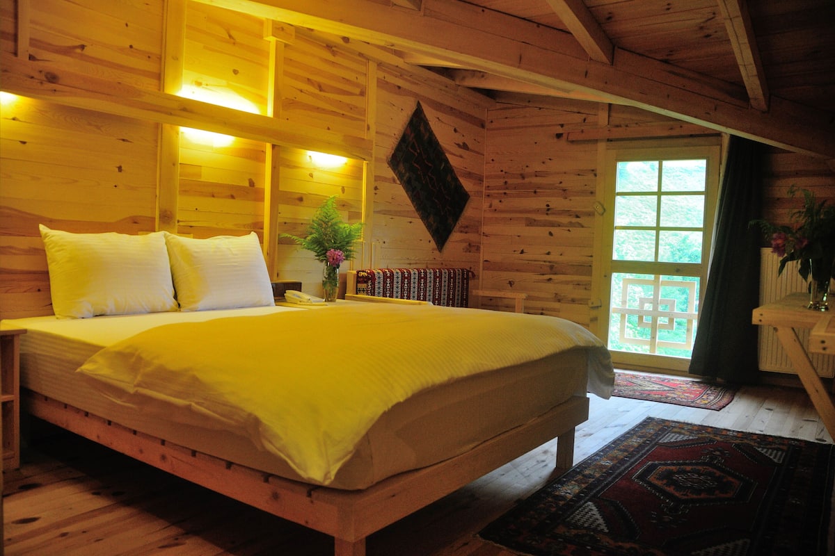 Quarto de cabana de madeira com uma cama, uma mesa de cabeceira, um vaso com flores e uma janela com vista para o verde.