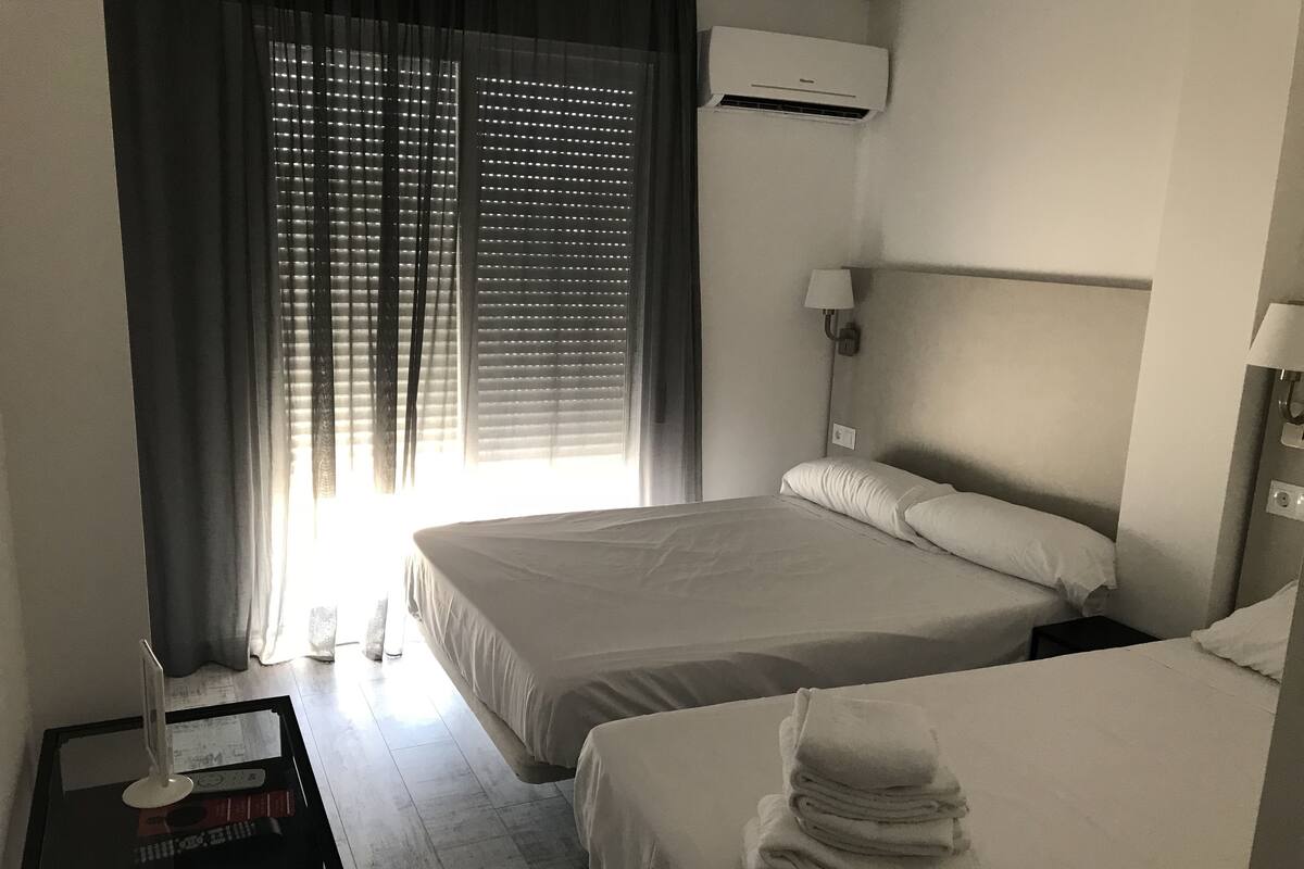 Ein Hotelzimmer mit zwei Betten, einem Nachttisch mit Lampe, einem Fenster mit Jalousien und einer Klimaanlage.