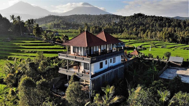 Une maison à deux étages avec un toit rouge, entourée de champs verdoyants et avec une montagne en arrière-plan.