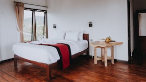 Un lit bien fait, avec du linge de lit blanc et un élément décoratif rouge, une table en bois sur laquelle sont disposés des fruits, et une vue sur la verdure par la fenêtre.