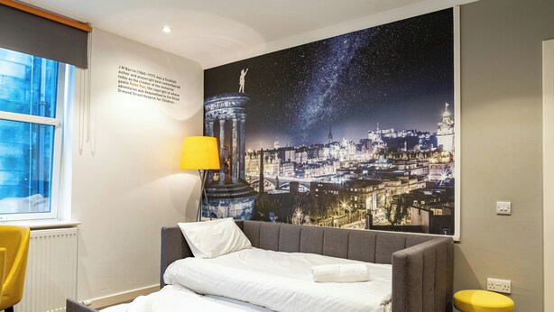 A hotel room with a bed, a window, a lamp, a stool, and a poster of a cityscape. - Stewart Aparthotel Edinburgh (Edinburgh)