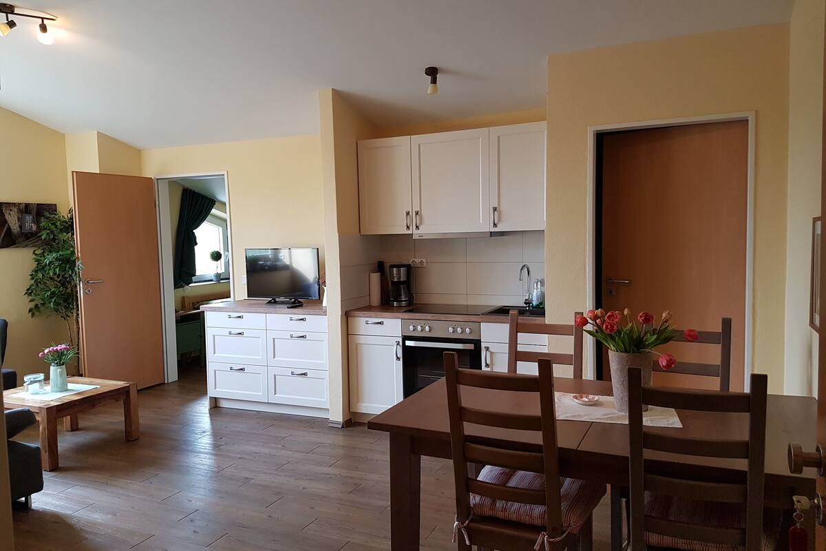 Uma cozinha compacta com armários brancos, fogão e pia. Há uma mesa de jantar com cadeiras e um vaso de planta.