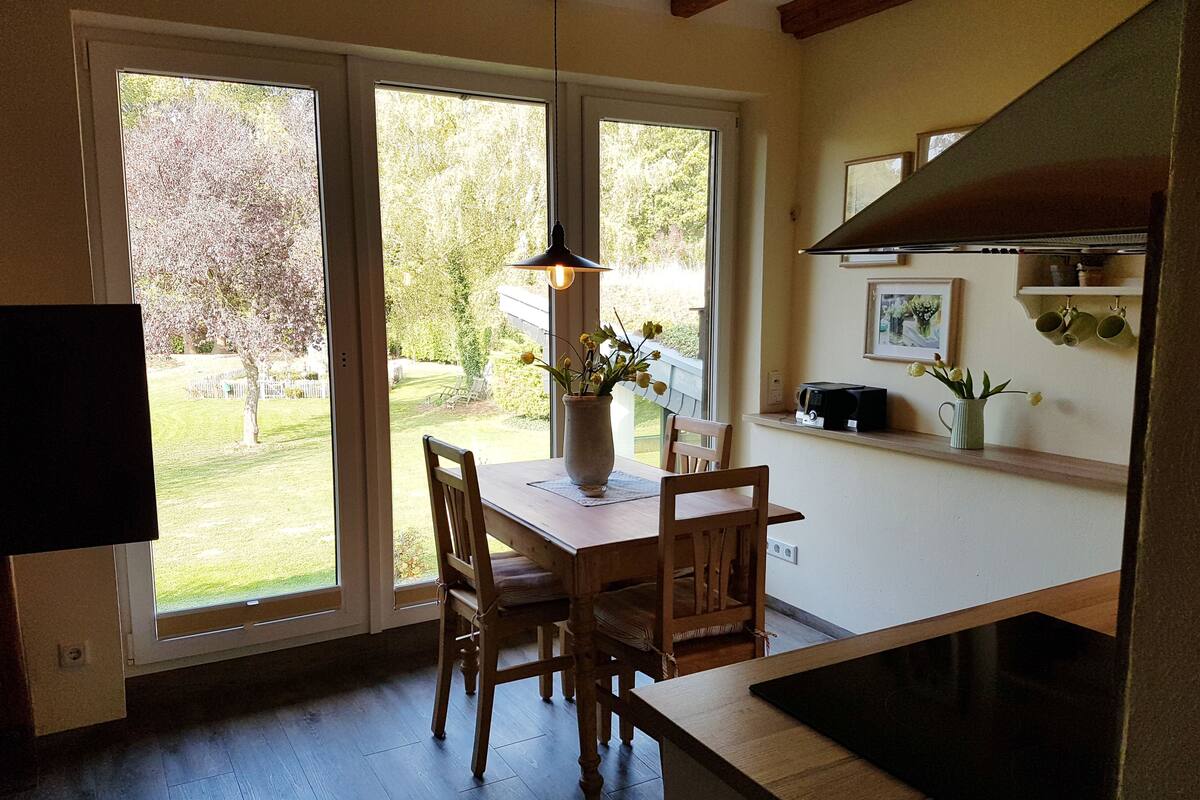 Uma área de jantar com mesa e cadeiras de madeira, um vaso com flores e vista para um jardim através de grandes janelas.