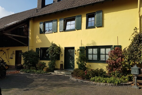 Un edificio amarillo con contraventanas verdes, una puerta verde y un pequeño jardín con diversas plantas.