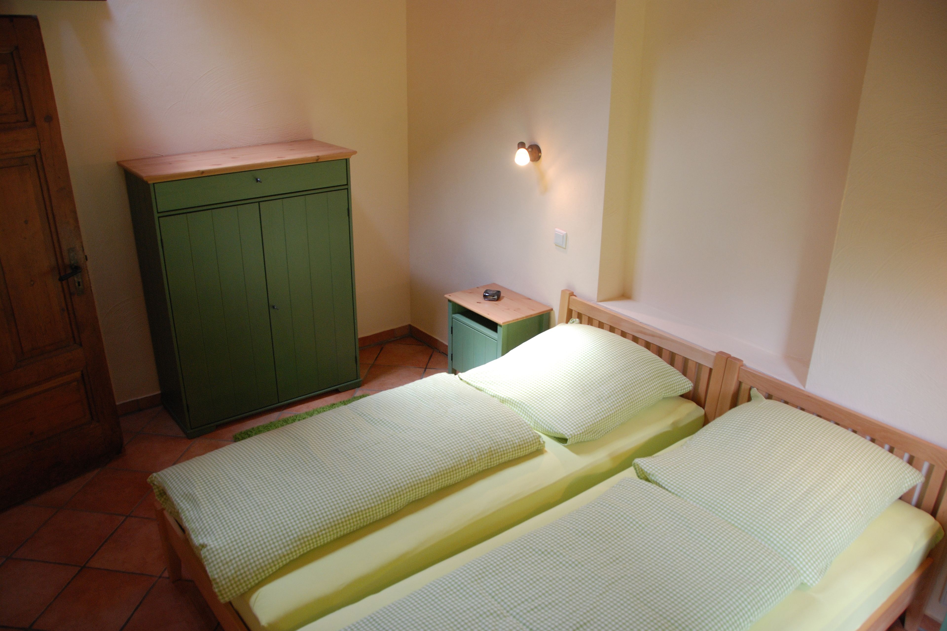 Une petite pièce unique comprenant deux lits, une armoire en bois, une petite table et un luminaire fixé au mur.