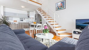 Ett modernt vardagsrum med en grå soffa, ett litet runt bord med en växt, en trappa som leder upp till en övre våning och ett kök i bakgrunden.