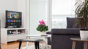 Ett modernt vardagsrum med en platt-TV, en grå soffa, ett litet runt bord, ett vitt soffbord, en krukväxt och en bokhylla.