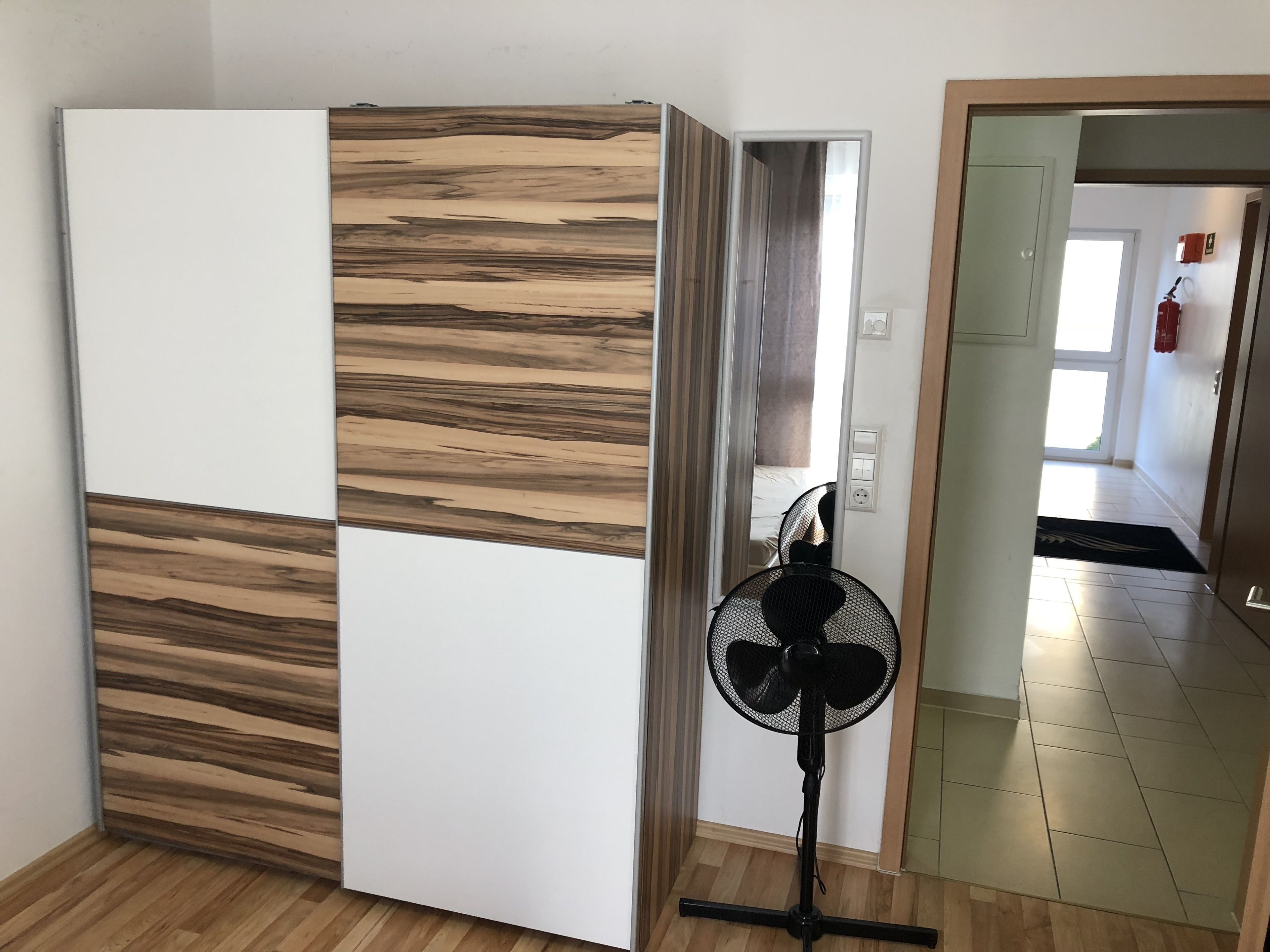 Una habitación de diseño moderno, con un mueble blanco grande con textura de madera, un ventilador de pie y vista a la habitación contigua.