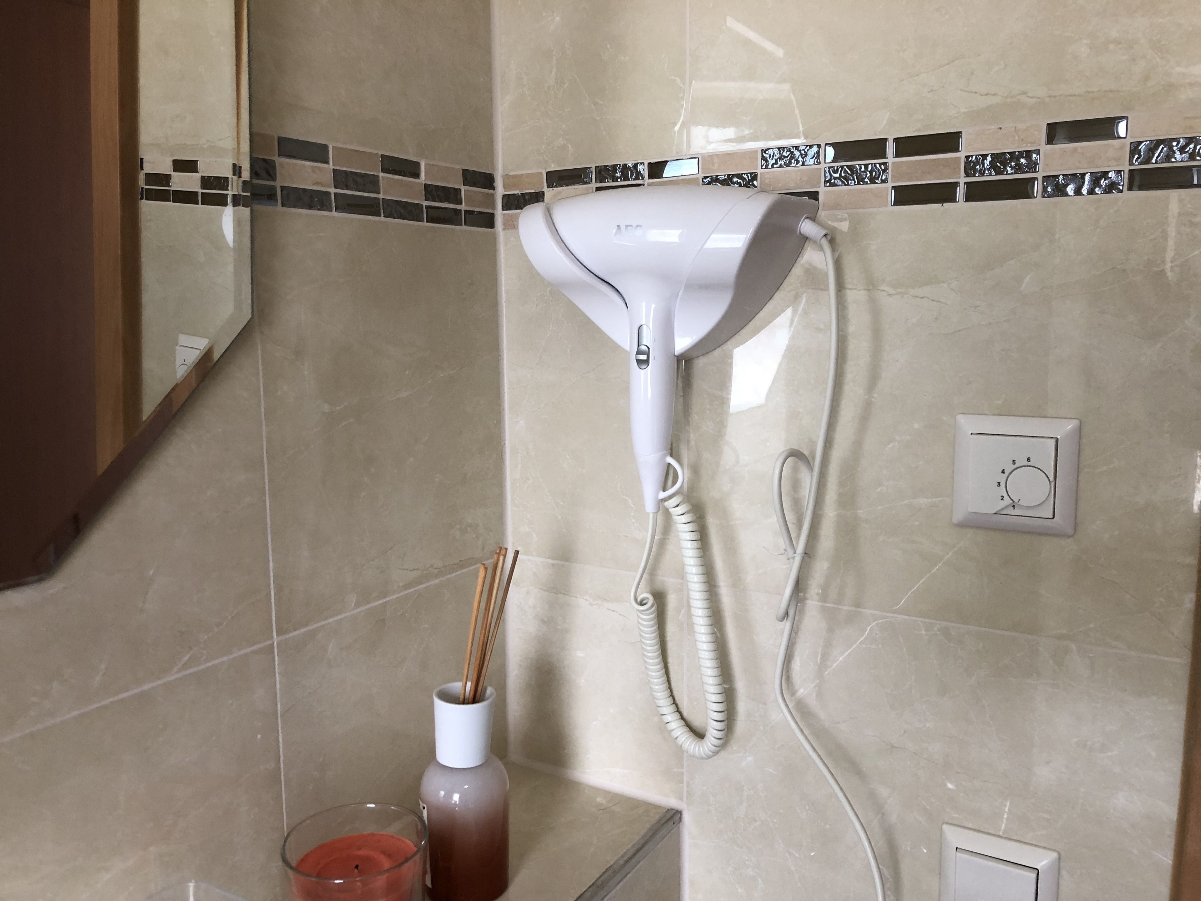Un baño con un secador de pelo montado en la pared, un difusor, una vela y un pequeño florero con cañas de juncos.