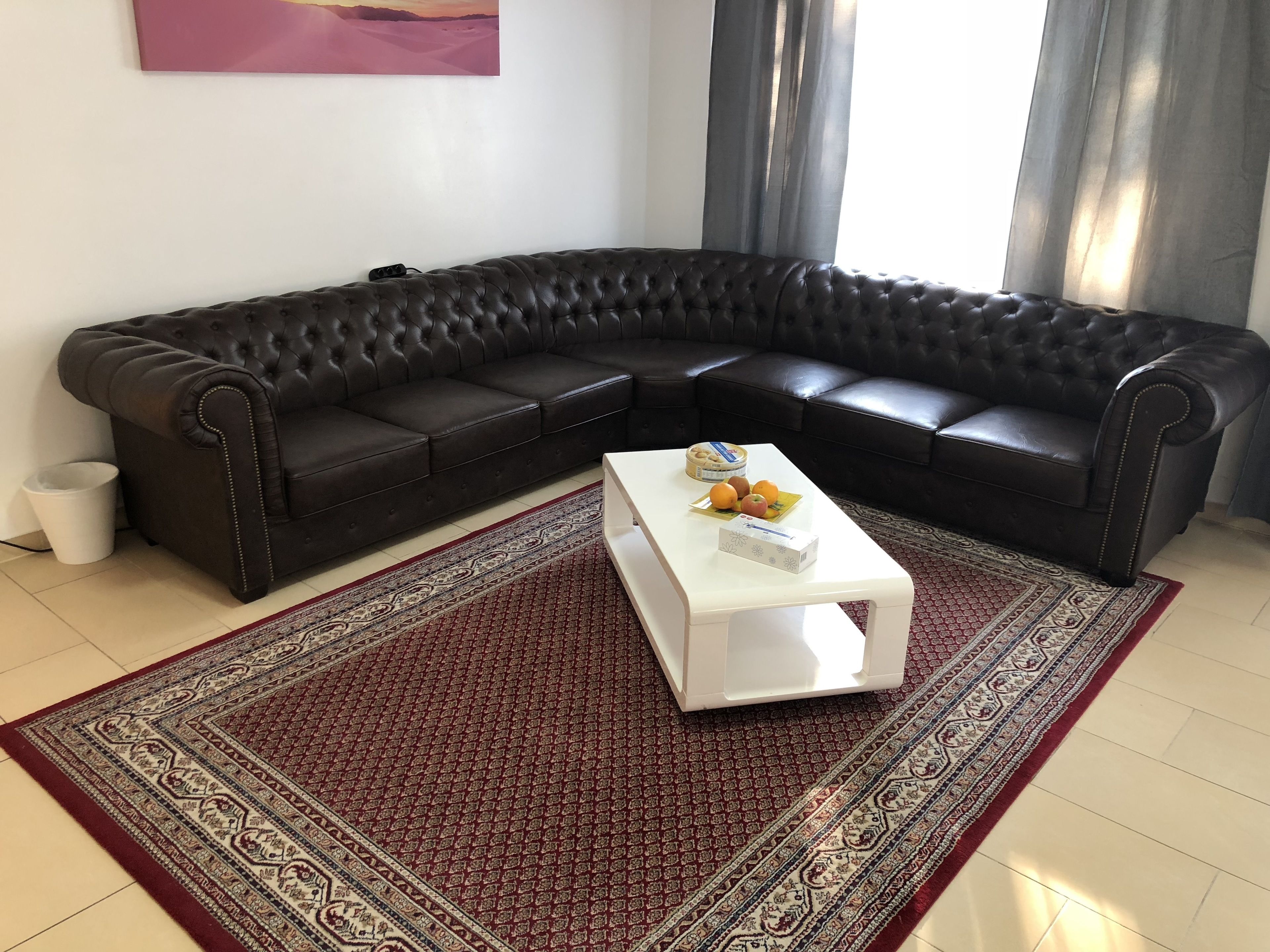 Een moderne woonkamer met een zwarte leren hoekbank, een witte salontafel met fruit, en een kamerkleed met een patroon.