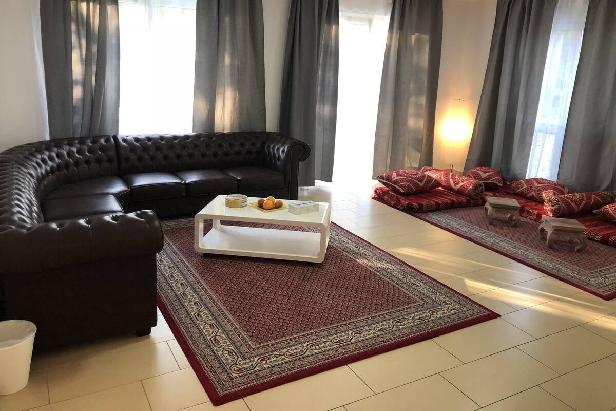 Un salon avec un canapé en cuir marron foncé, une table basse blanche ornée de fruits, un tapis à motifs et des coussins rouges.