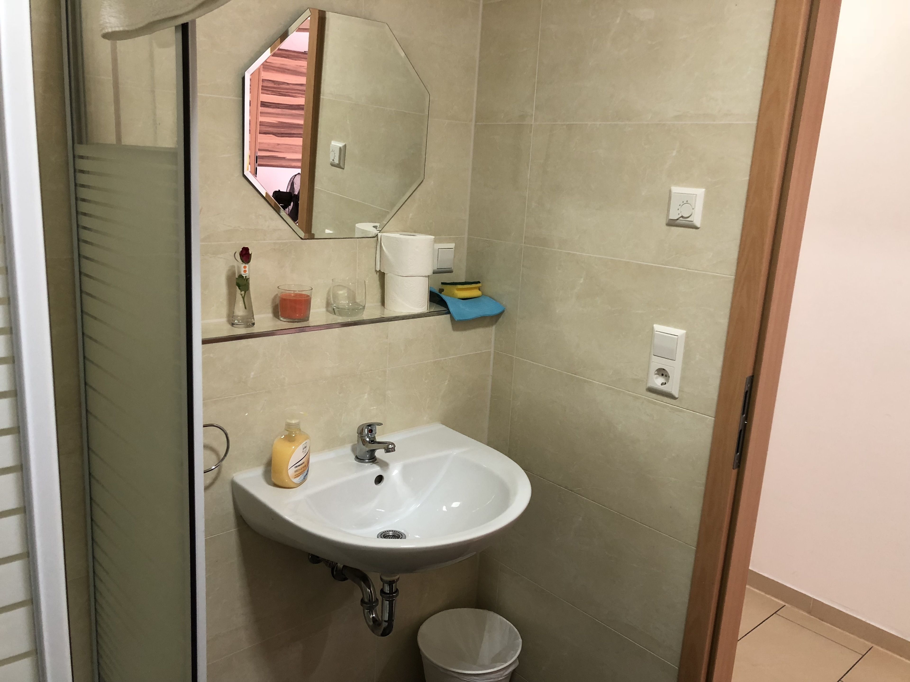 Un baño con un lavabo blanco, un espejo, un perchero para toallas, un cubo de basura pequeño y un estante con artículos de tocador.