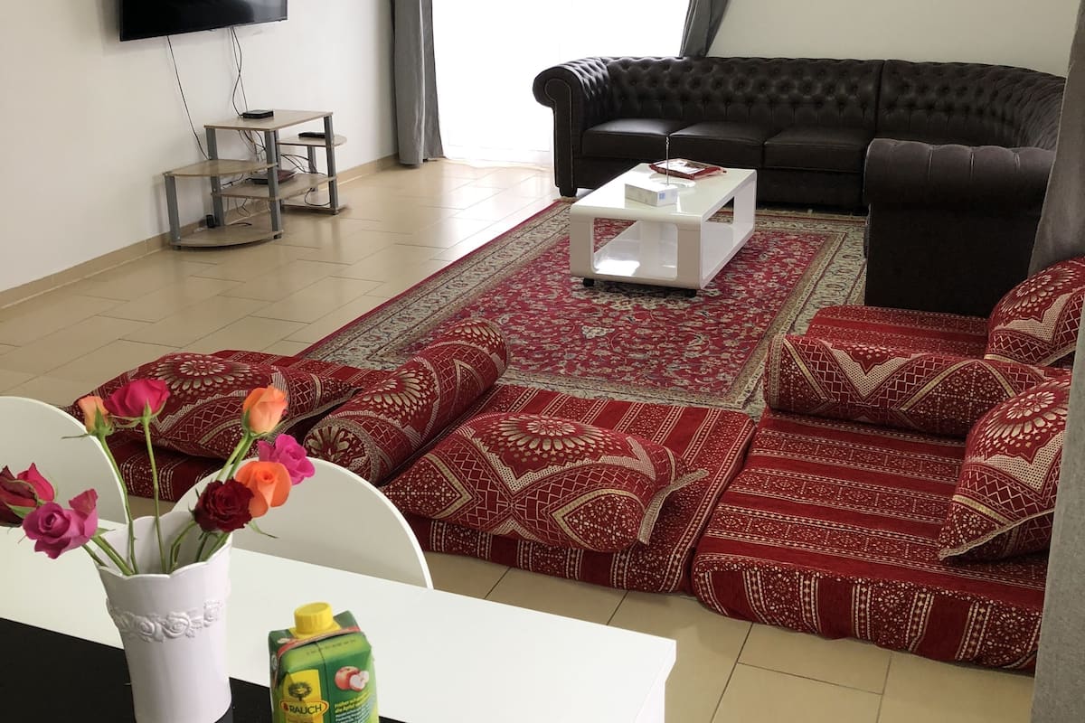 Un salon avec un téléviseur à écran plat fixé au mur, un canapé en cuir noir, un tapis rouge à motifs et une table basse blanche.