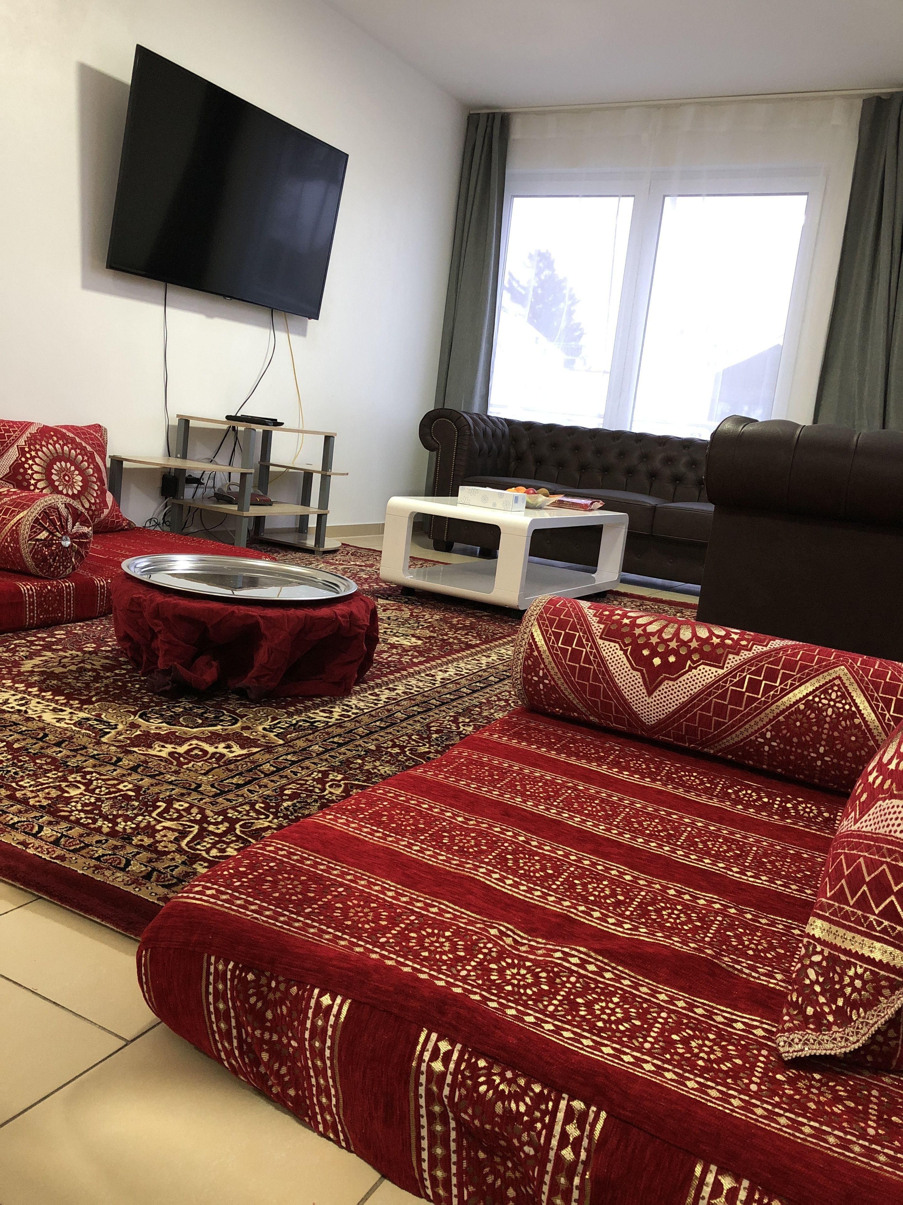 Sala de estar com televisão de tela plana na parede, sofá marrom escuro, tapete vermelho estampado e mesa lateral branca com um controle remoto.