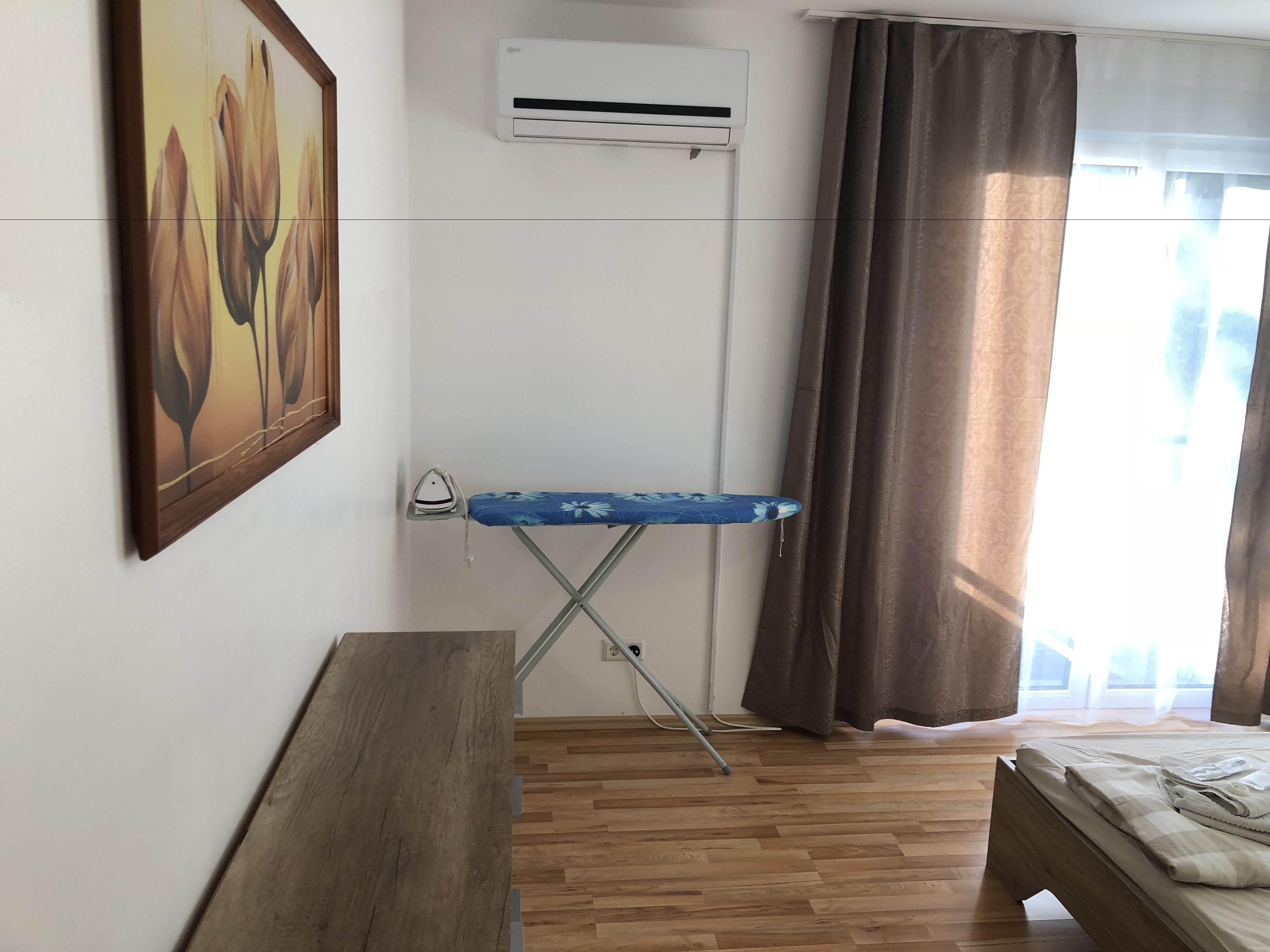 A room with a wooden floor, a bed with a white blanket, a blue ironing board, an air conditioner, and a painting on the wall.
