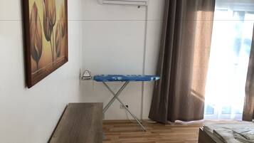 A room with a wooden floor, a bed with a white blanket, a blue ironing board, an air conditioner, and a painting on the wall.