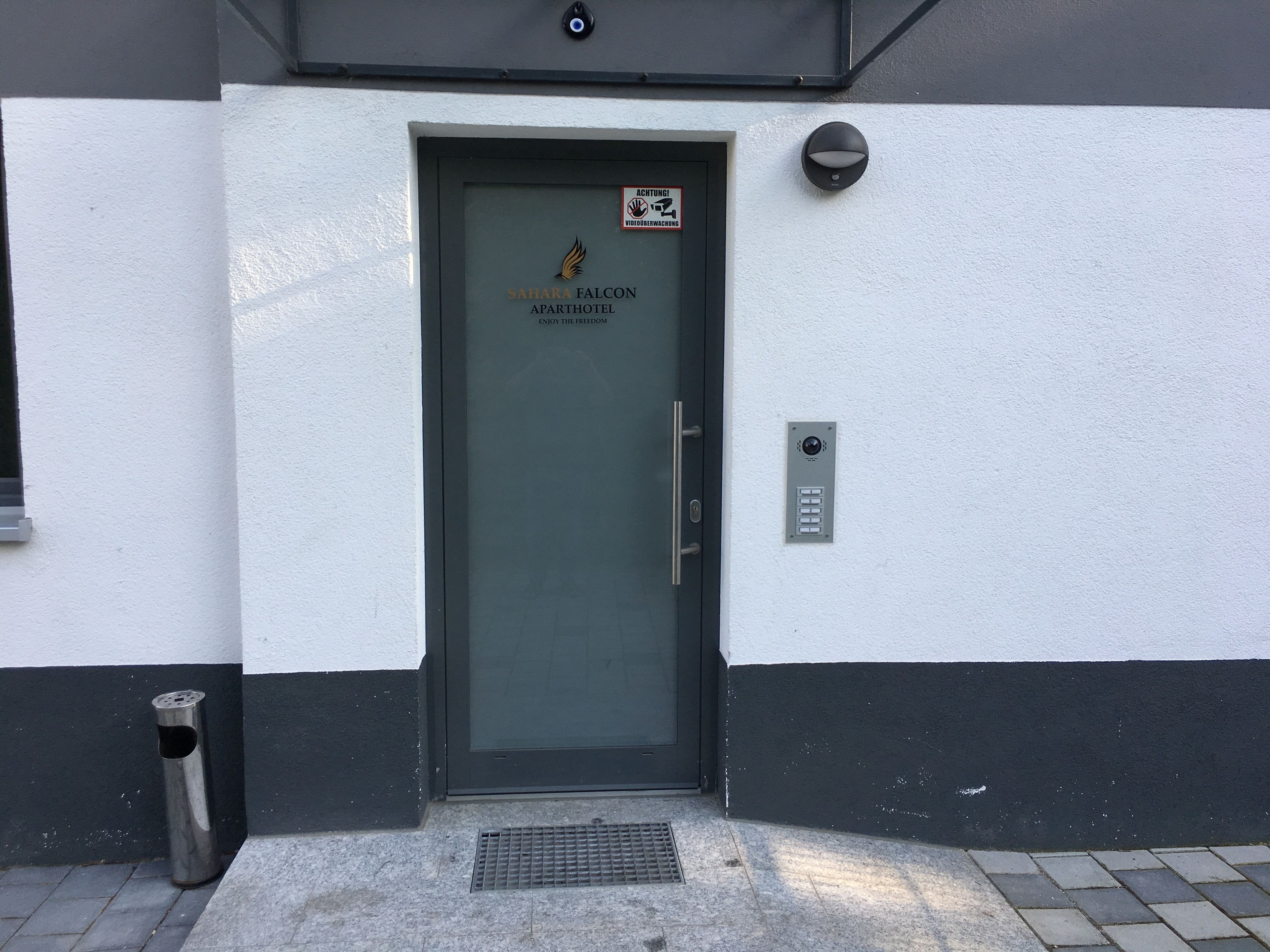 A door with a sign that reads "SAHARA FALCON APARTHOTEL ENJOY THE FREEDOM" and a security camera above it.