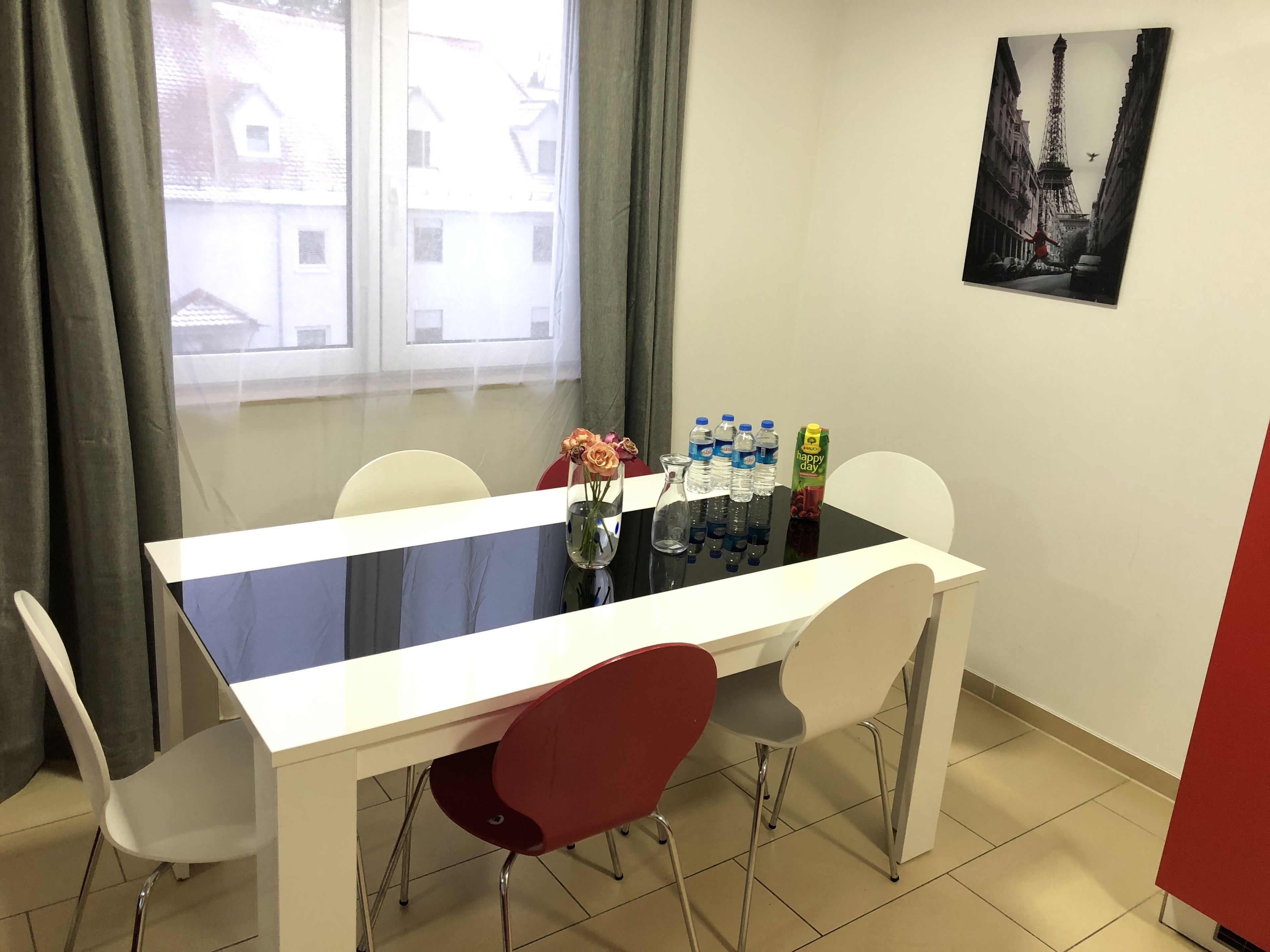 Un comedor con una mesa de vidrio, sillas blancas y una silla roja. Hay botellas de agua y un cartón de jugo sobre la mesa. Se ve una ventana con cortinas, y en la pared hay un cuadro con la Torre Eiffel.