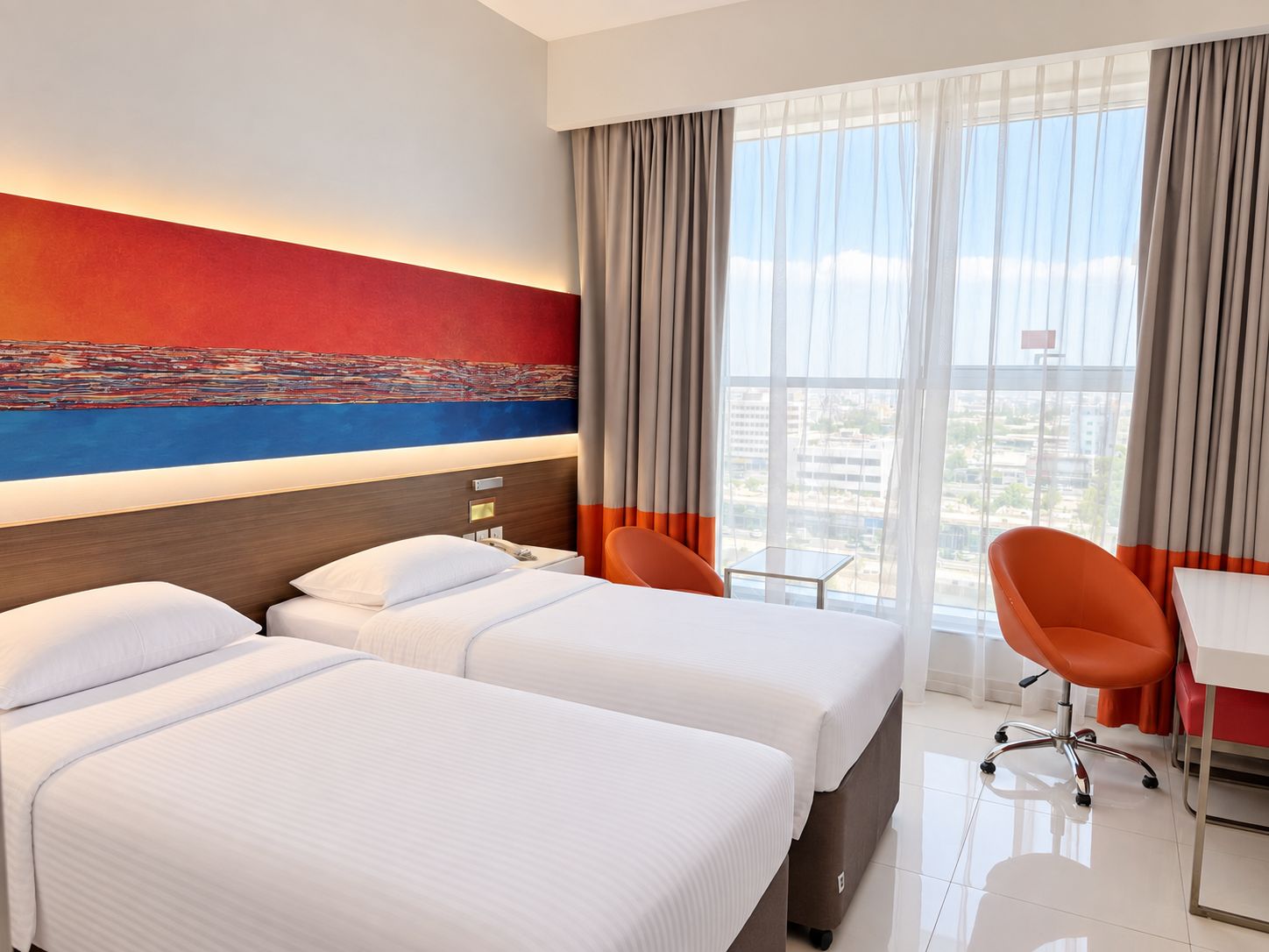 Une chambre d’hôtel avec deux lits, un bureau et une chaise. Il y a une grande fenêtre avec des rideaux, et un tableau au mur.