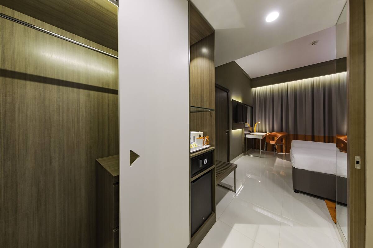 A modern hotel room with a wooden wardrobe, a small kitchenette, a bed, a desk, and a TV.