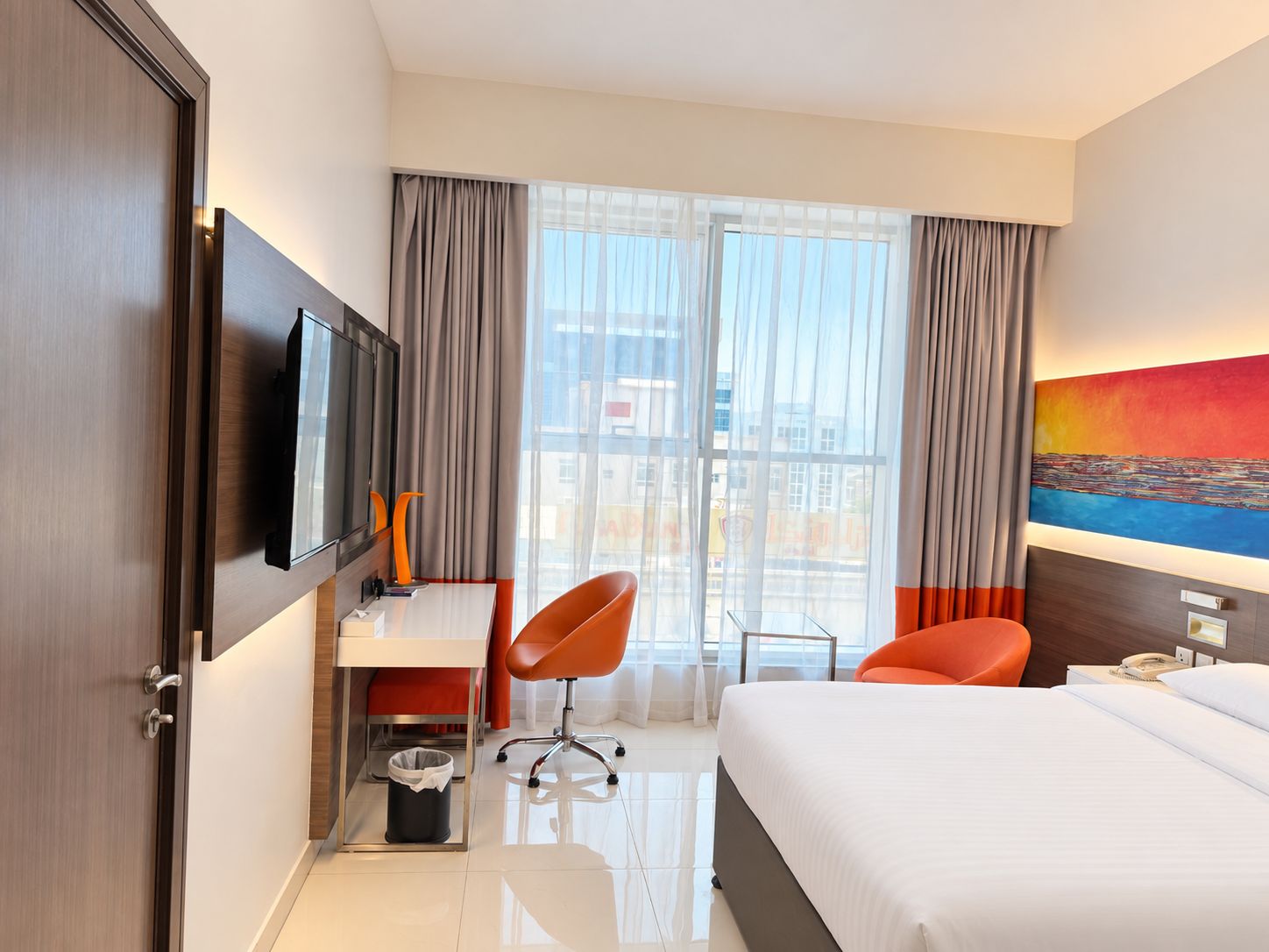 A hotel room with a bed, a desk, a chair, a TV, and a painting on the wall.