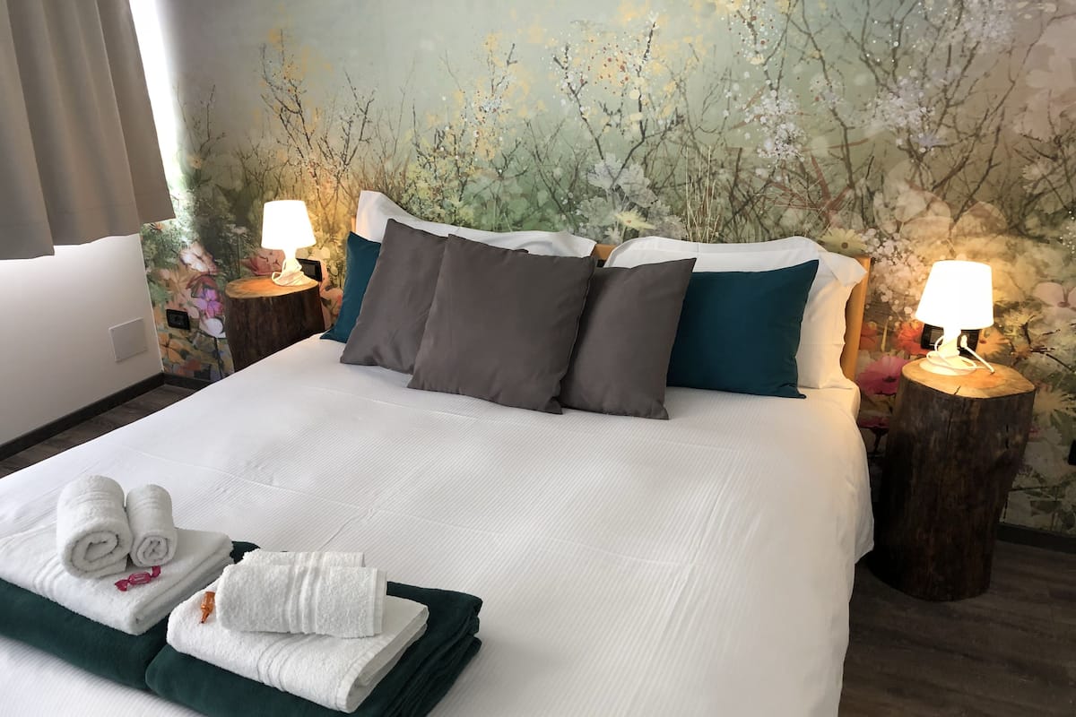 A neatly made bed with white and dark green linens, two pillows, and a stack of folded towels. There are two bedside tables with lamps and a floral wall mural.