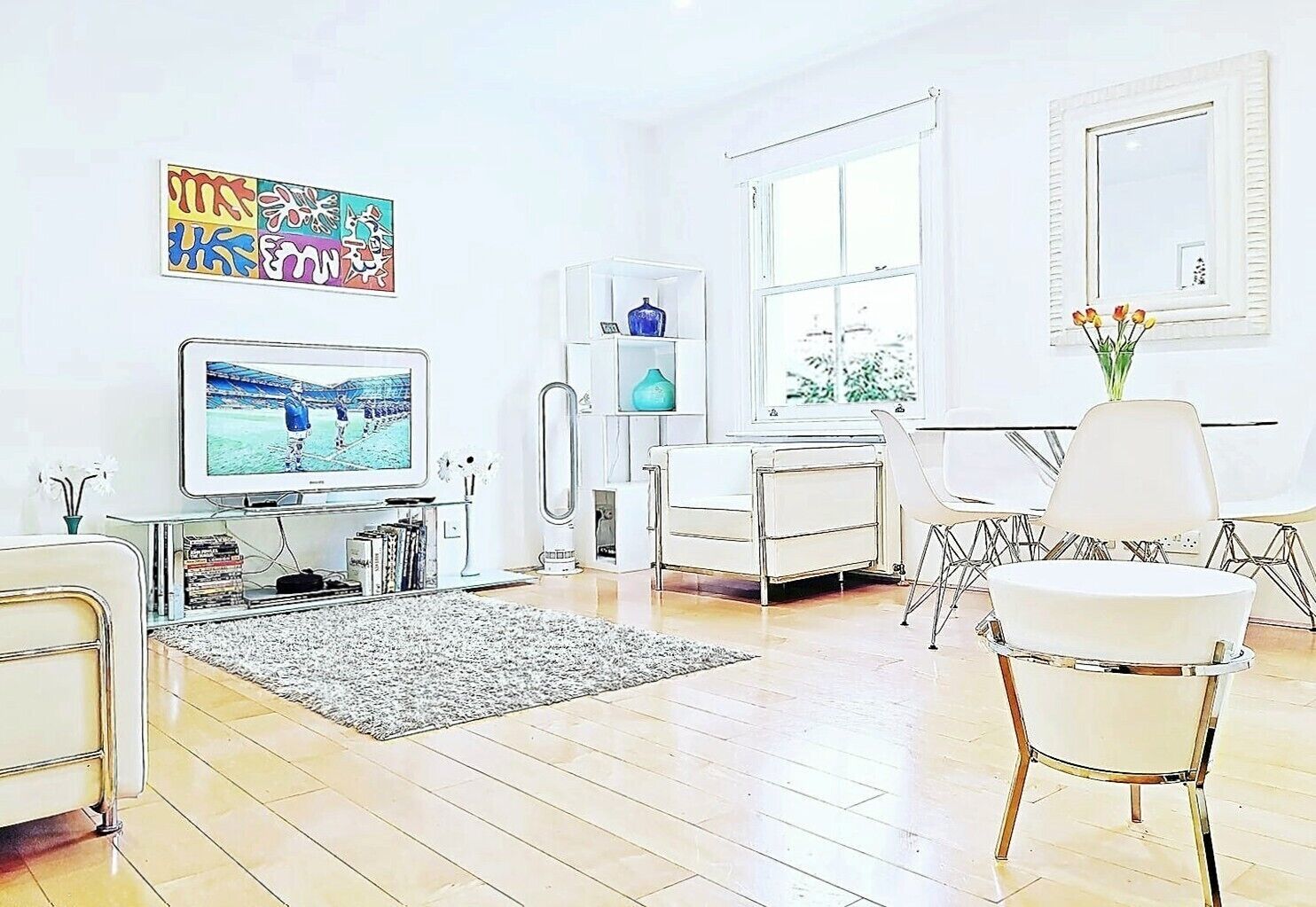 Ein modernes Wohnzimmer mit Fernseher, weißen Möbeln, einem Glastisch und einem großen Fenster.