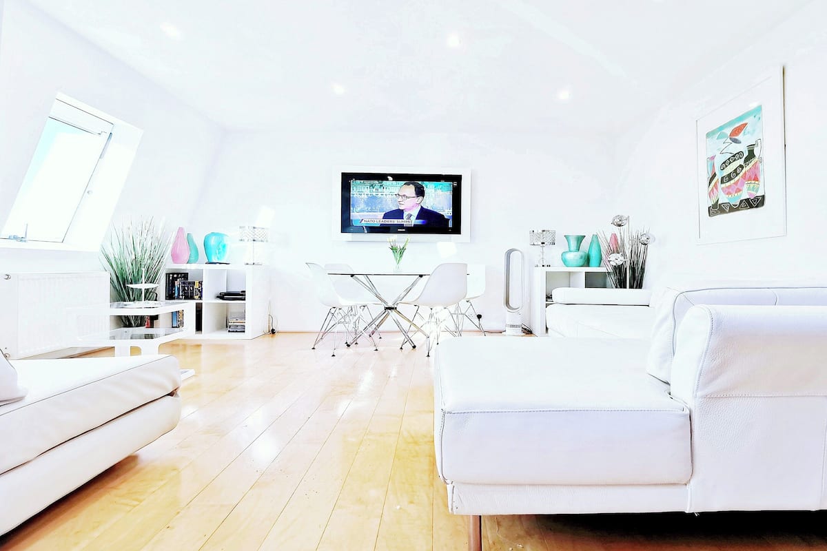 A modern living room with a flat-screen TV, white sofas, a glass coffee table, and a dining area with chairs and a small table.
