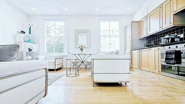 A modern kitchen with wooden cabinets, a dining area with a white table and chairs, and a large window providing natural light. - Luxury Designer Apartments Hammersmith 1 (London)