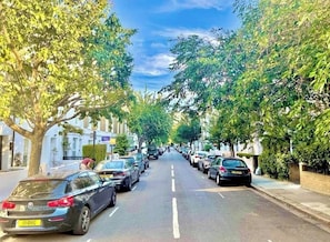 Une rue bordée d’arbres, avec des voitures garées de part et d’autre et des immeubles qui la bordent.