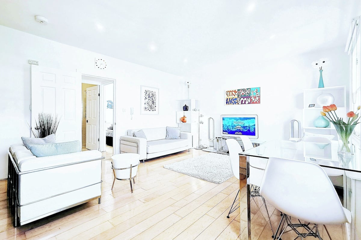 A modern living room with a white sofa, a dining table with chairs, a TV, and artwork on the walls.