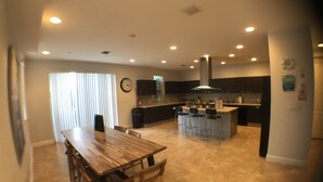 A modern kitchen with dark cabinets, a central island with bar stools, and a dining area with a wooden table and chairs. - Private Home by Ideal Experience VR (Fort Lauderdale)