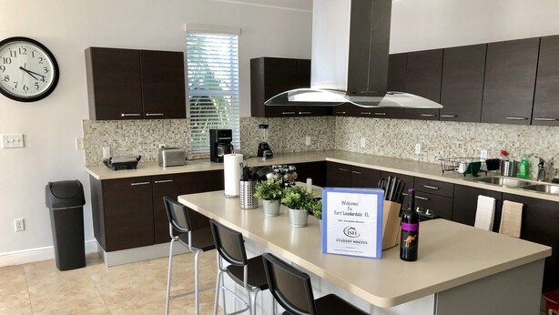 A modern kitchen with dark cabinets, a central island with bar stools, a clock on the wall, and a welcome sign for ISH International Student Houses. - Private Home by Ideal Experience VR (Fort Lauderdale)