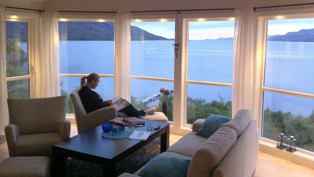 A room with large windows offering a view of a lake and mountains. There is a sofa and a chair, a table with a laptop and papers.