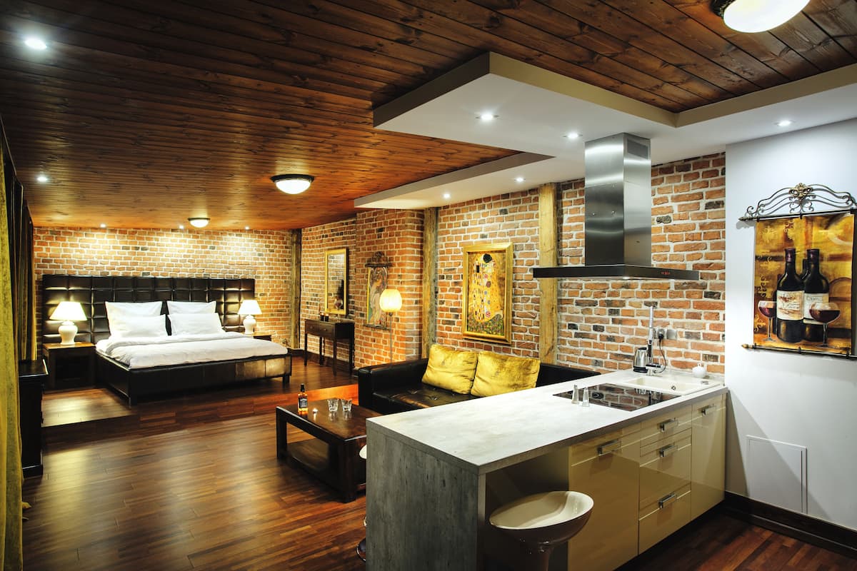 Une chambre d’hôtel moderne avec un mur en briques, un plafond en bois, un lit, un canapé, une table basse et un coin cuisine équipé d’un évier et de placards.